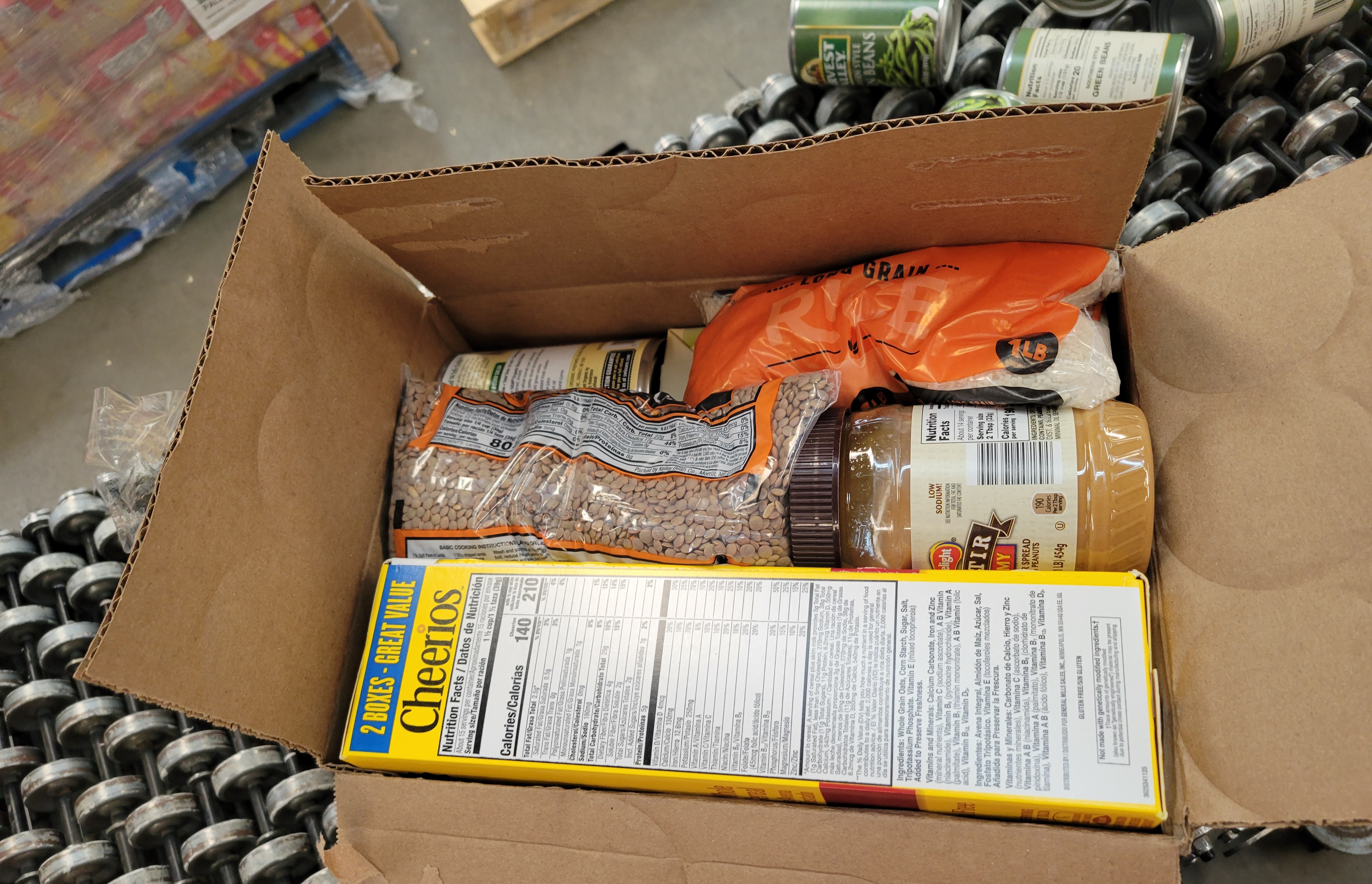 Box of dry goods on a conveyor belt with cans of green beans behind it.